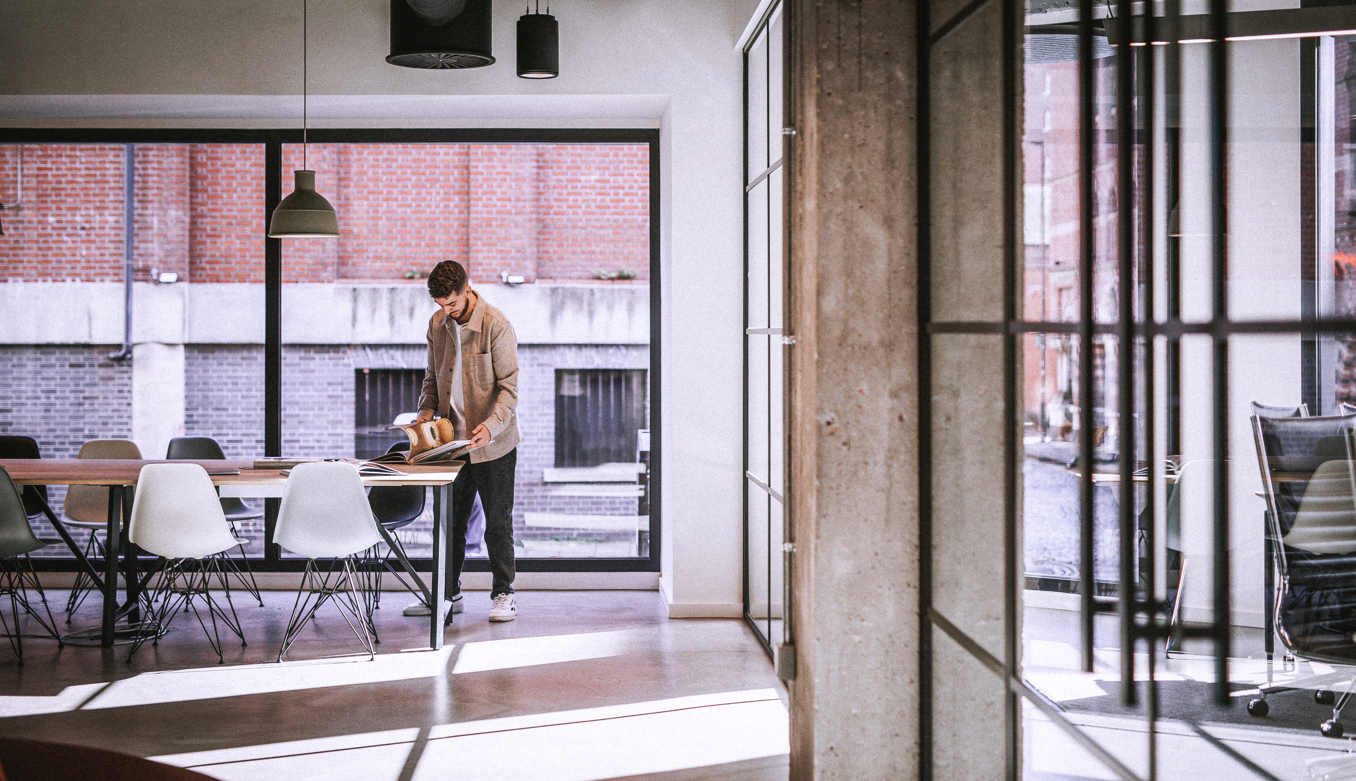 A spacious industrial-style interior with exposed brick walls, large windows, and natural light pouring in during sunset. Wooden desks with plants and workspace materials are arranged along the walls, with a view of greenery and an outdoor seating area.