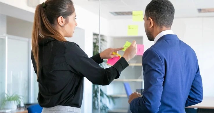 Duas pessoas escrevendo em post-its num quadro de vidro, vistas de costas