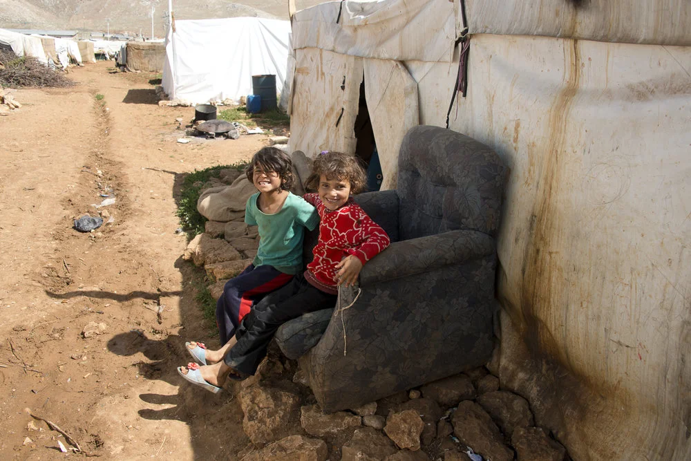 Two young children sitting on an old armchair outside a tent in a refugee camp