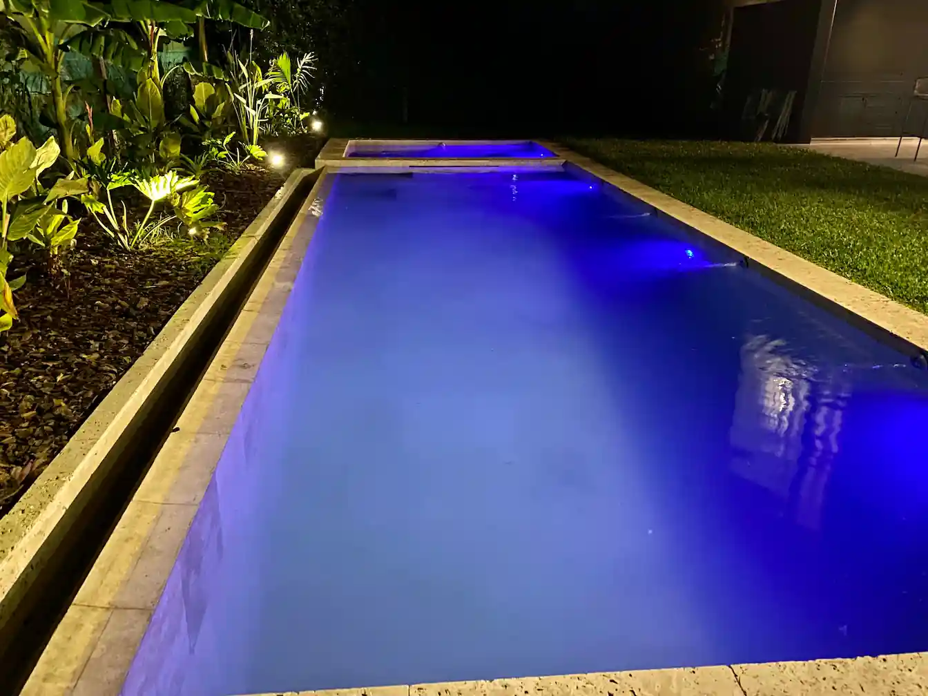 Piscina larga iluminada de noche con vibrantes luces LED azules, rodeada de un cantero decorativo con plantas tropicales iluminadas.