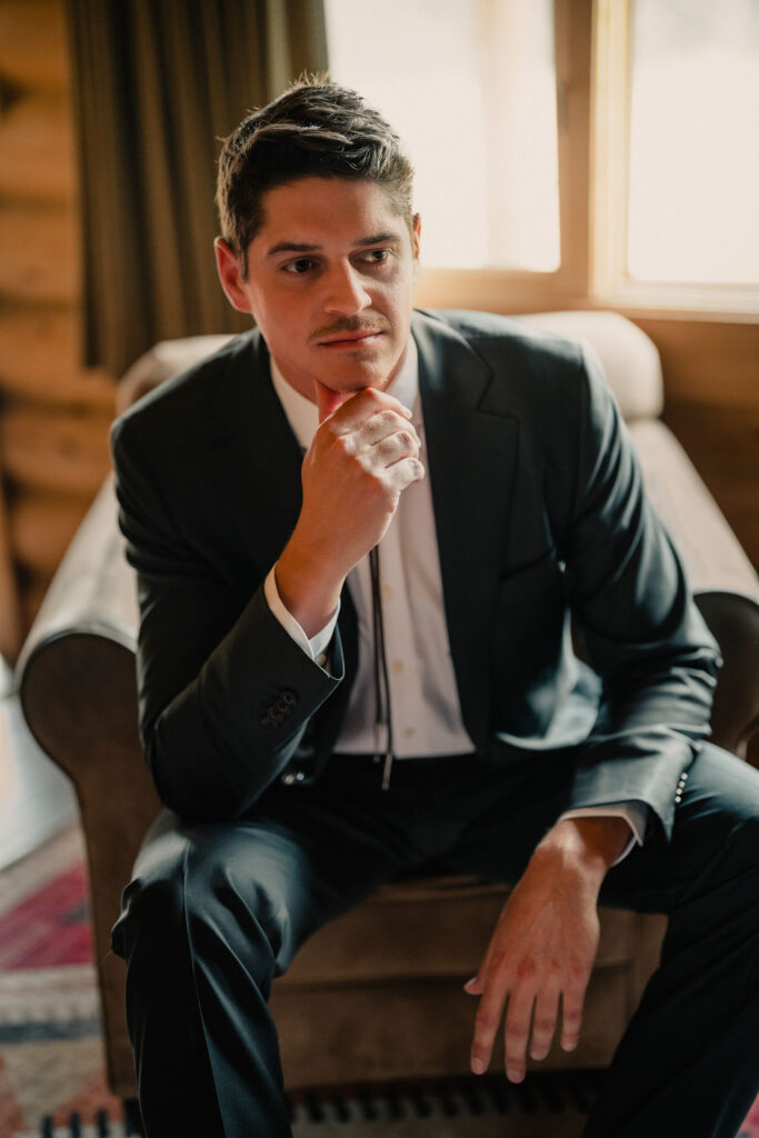 A portrait of the groom sitting in a leather armchair inside a log cabin, resting his chin on his hand and looking off-camera with a contemplative expression.
