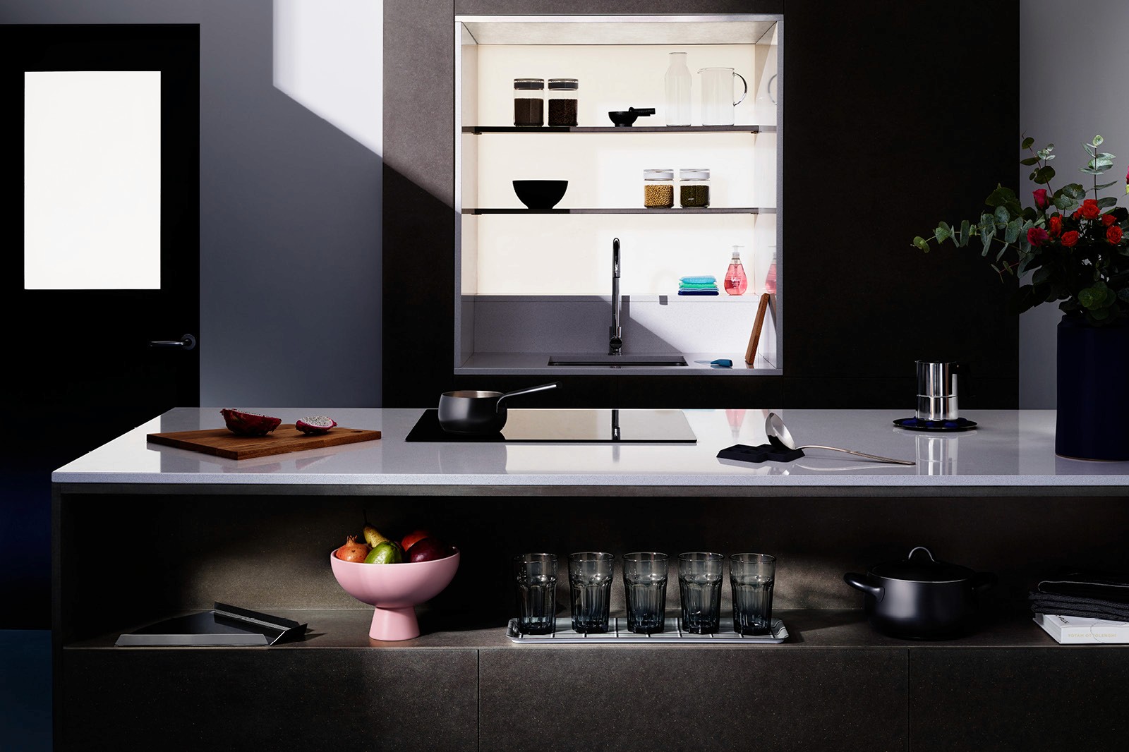 Modern kitchen interior with a sleek countertop, shelves displaying decor, and a large window letting in natural light.
