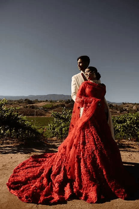 Modern Indian wedding portrait featuring a bride in a vibrant red lehenga and groom in a white sherwani, posed elegantly outdoors with a natural backdrop, reflecting contemporary wedding photography style.