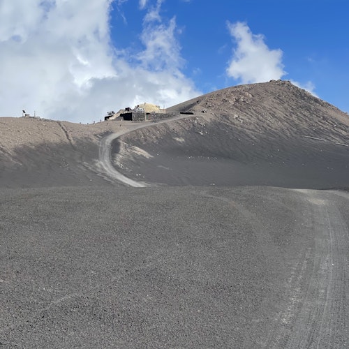 Um caminho de terra acidentado e sinuoso leva a edifícios no topo de uma encosta árida e rochosa sob um céu azul com nuvens esparsas.