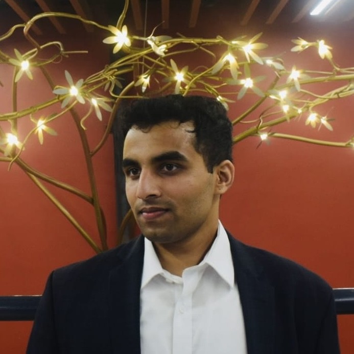 A young woman with long brown hair smiles warmly against a green background, dressed in a black jacket and white shirt.
