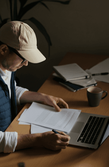 A person wearing a cap studies documents on a wooden table, with papers and a mug nearby, using a laptop.