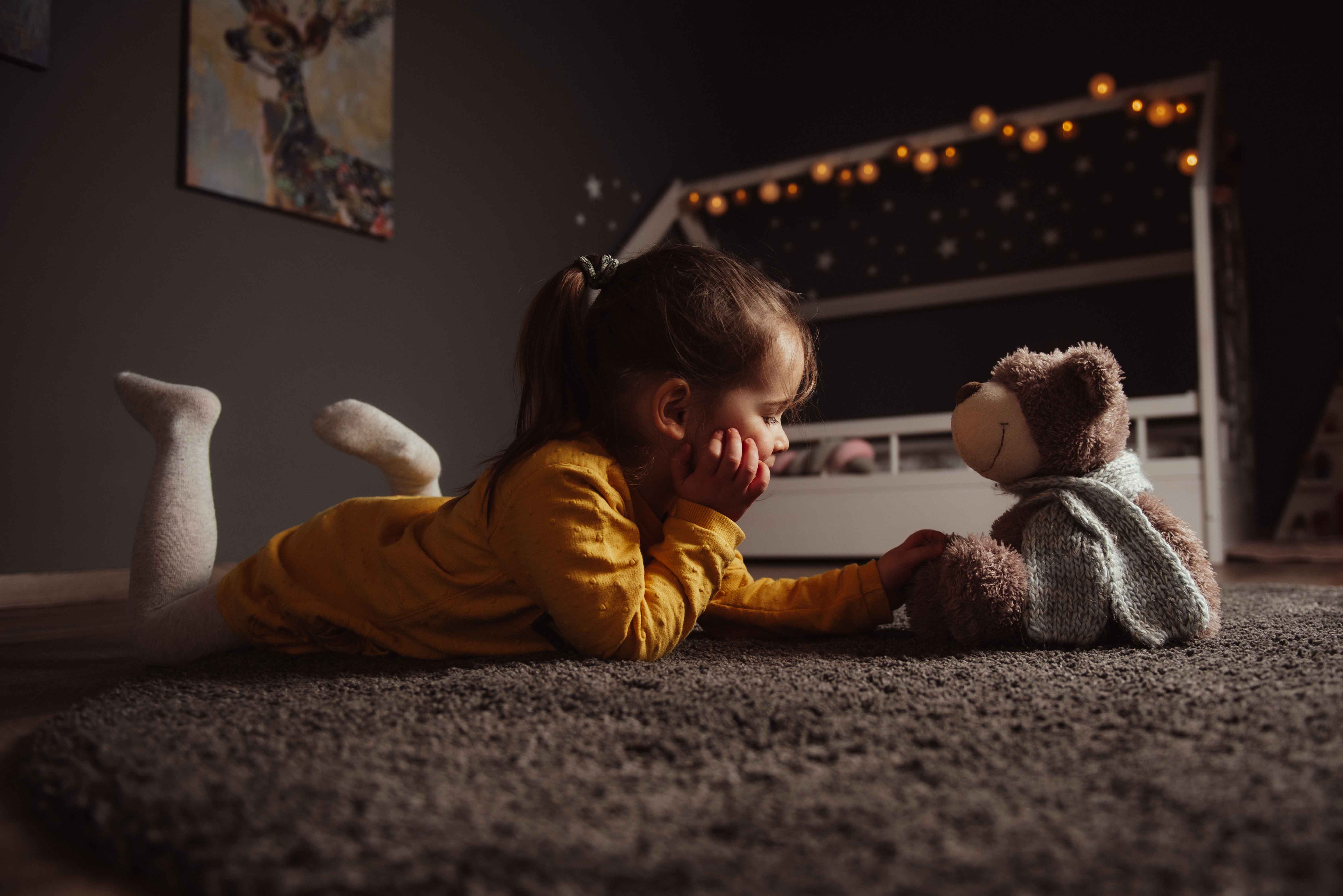 Kleines Mädchen in gelbem Kleid liegt auf Bauch auf Teppich und sieht Teddybär an, stimmungsvolle moderne Kitafotografie im modernen Kinderzimmer NRW, natürliches Licht.