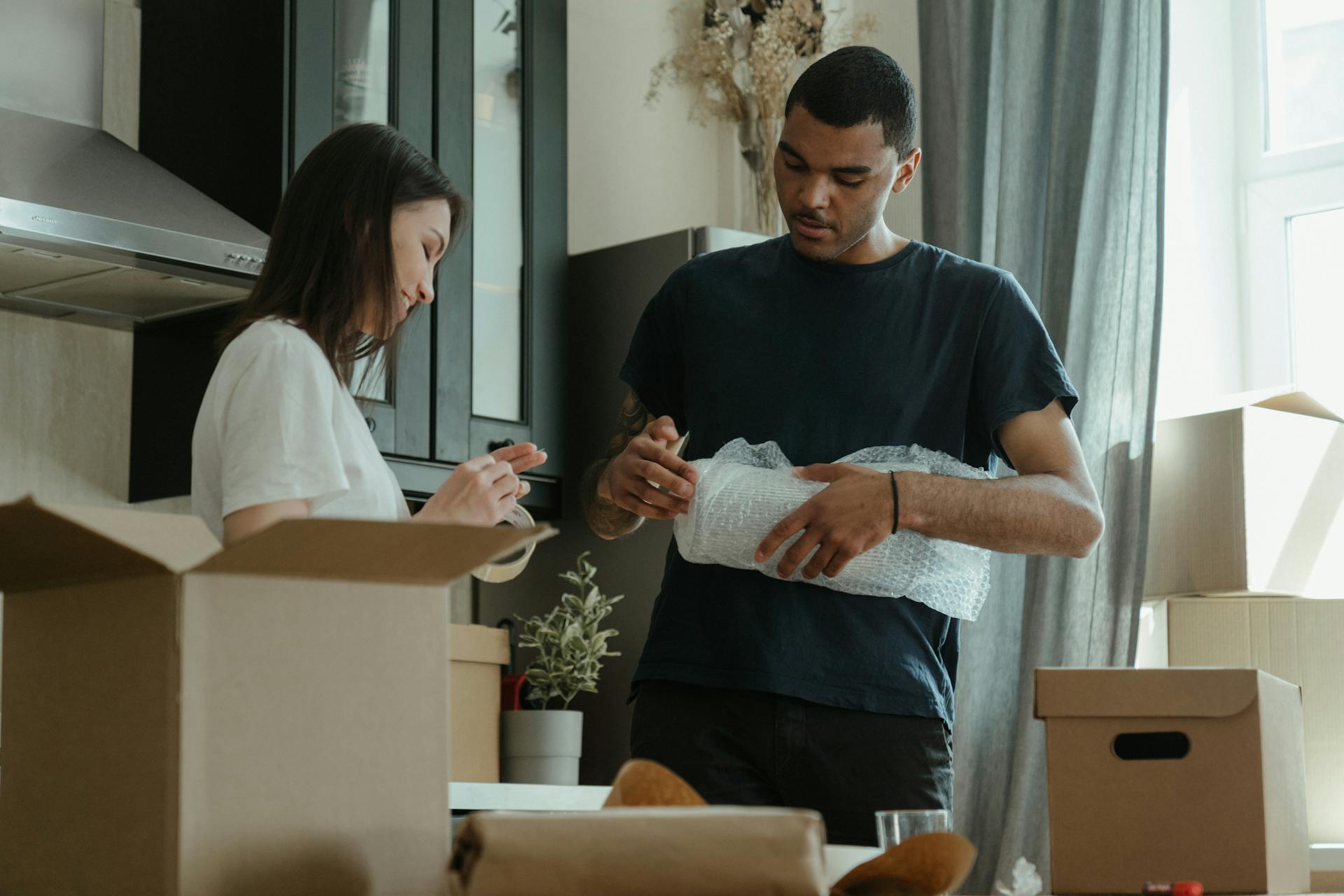 A couple stands inside, packing household items. A man with dark skin and short dark hair is carefully wrapping a cylindrical object in white bubble wrap, looking focused. A woman with dark hair pulled back is to his left, smiling slightly while sealing a cardboard box with tape.