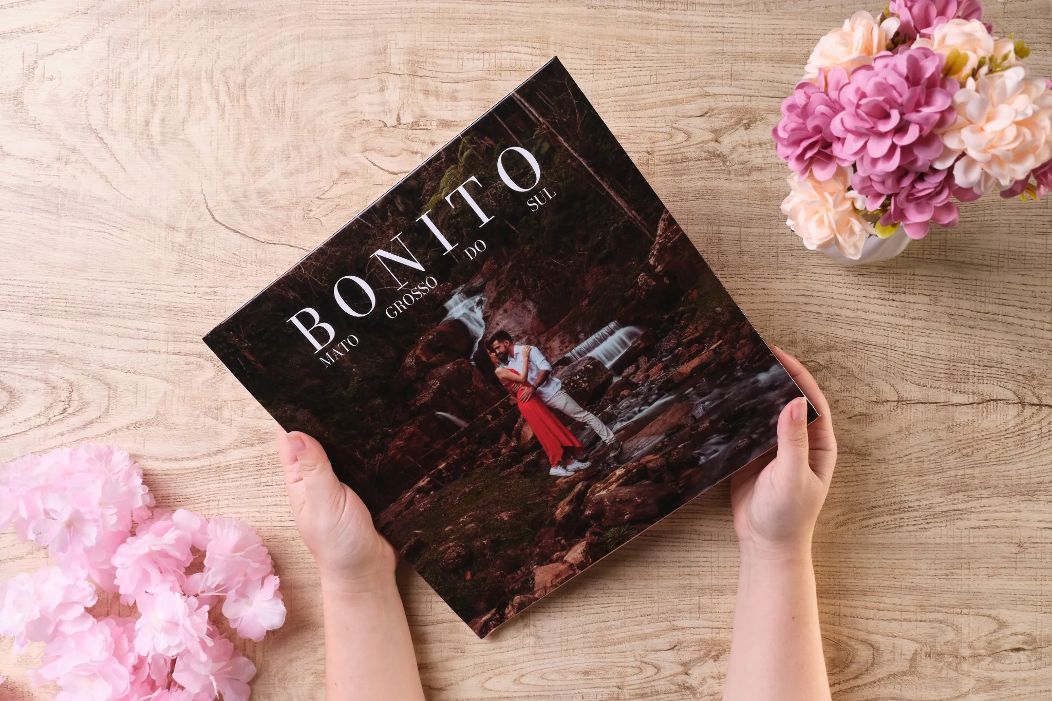 Mãos segurando fotolivro com capa de casal abraçado em cachoeira de Bonito, Mato Grosso do Sul, sobre mesa de madeira com flores decorativas