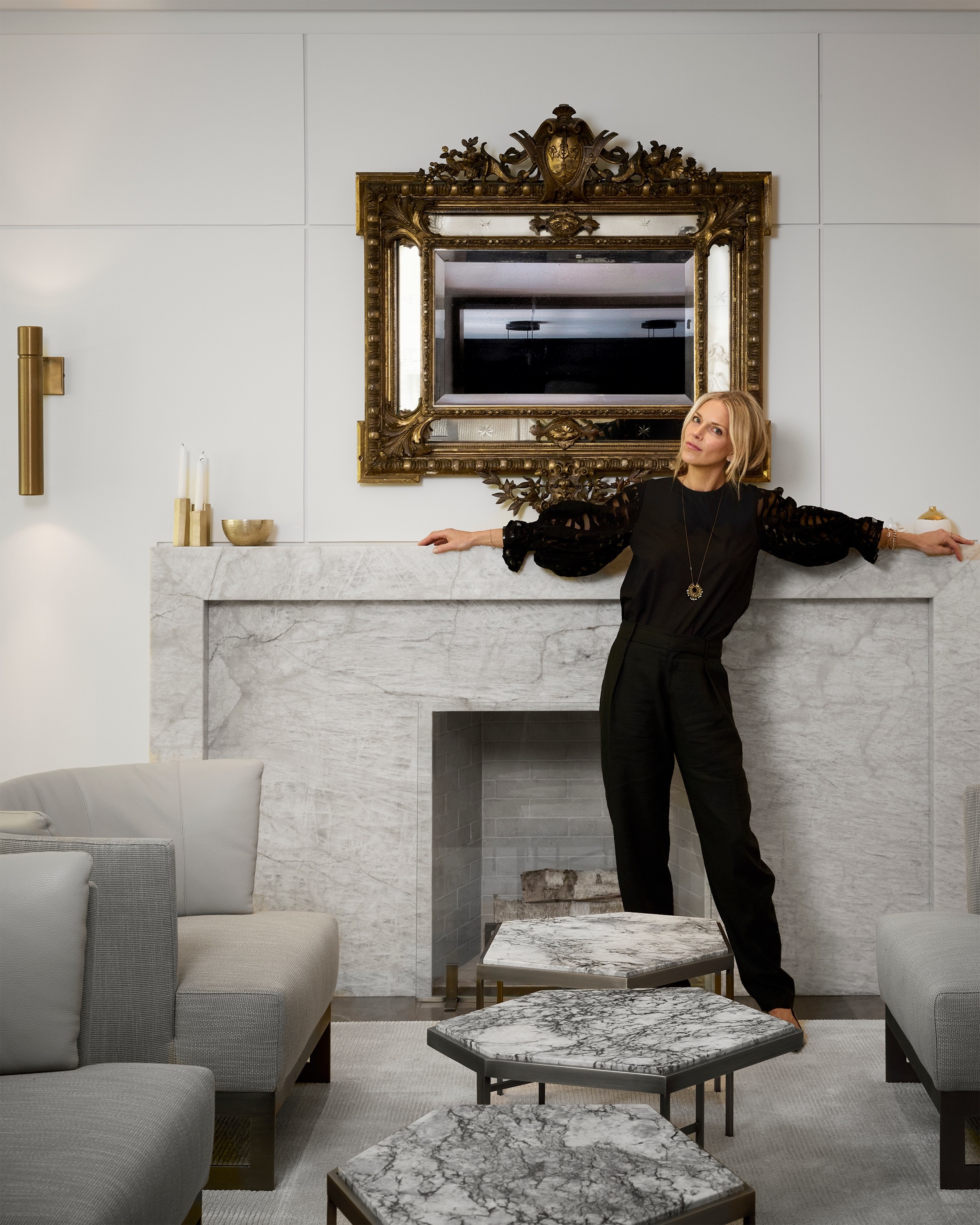 Kim Lambert, principal and creative director of Kim Lambert Design, leaning against a marble fireplace beneath an antique gilt mirror
