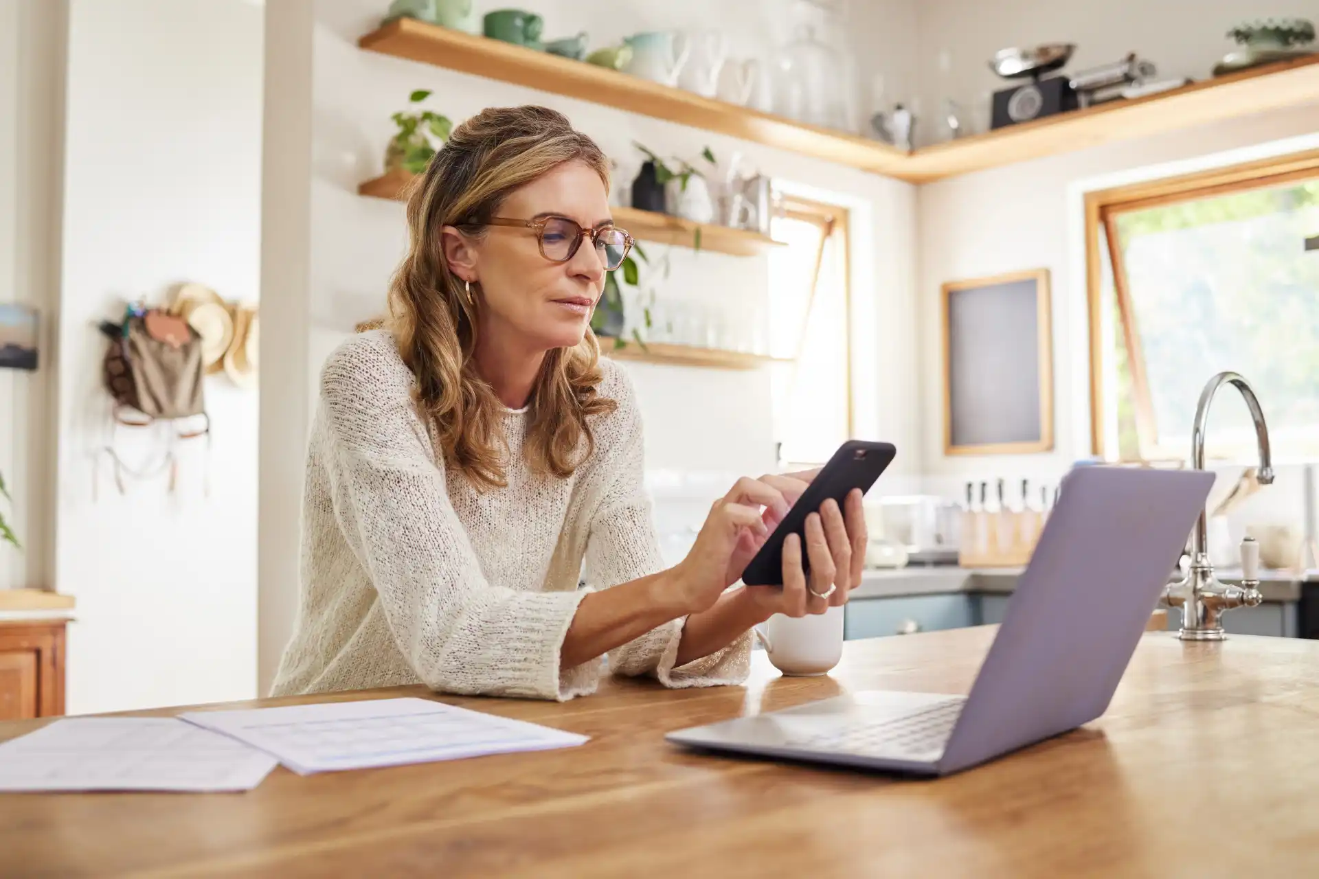 Een persoon zit aan een houten tafel met een smartphone in de hand en kijkt naar een laptop in een lichte, moderne keuken.