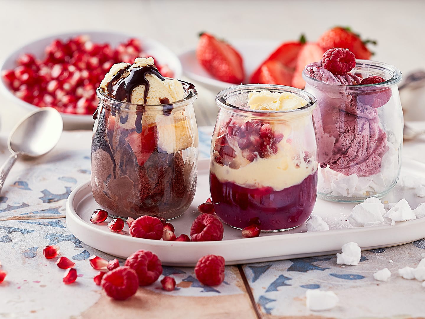 Three dessert jars, chocolate sundae, raspberry parfait, berry ice cream with fresh berries on tiled tray