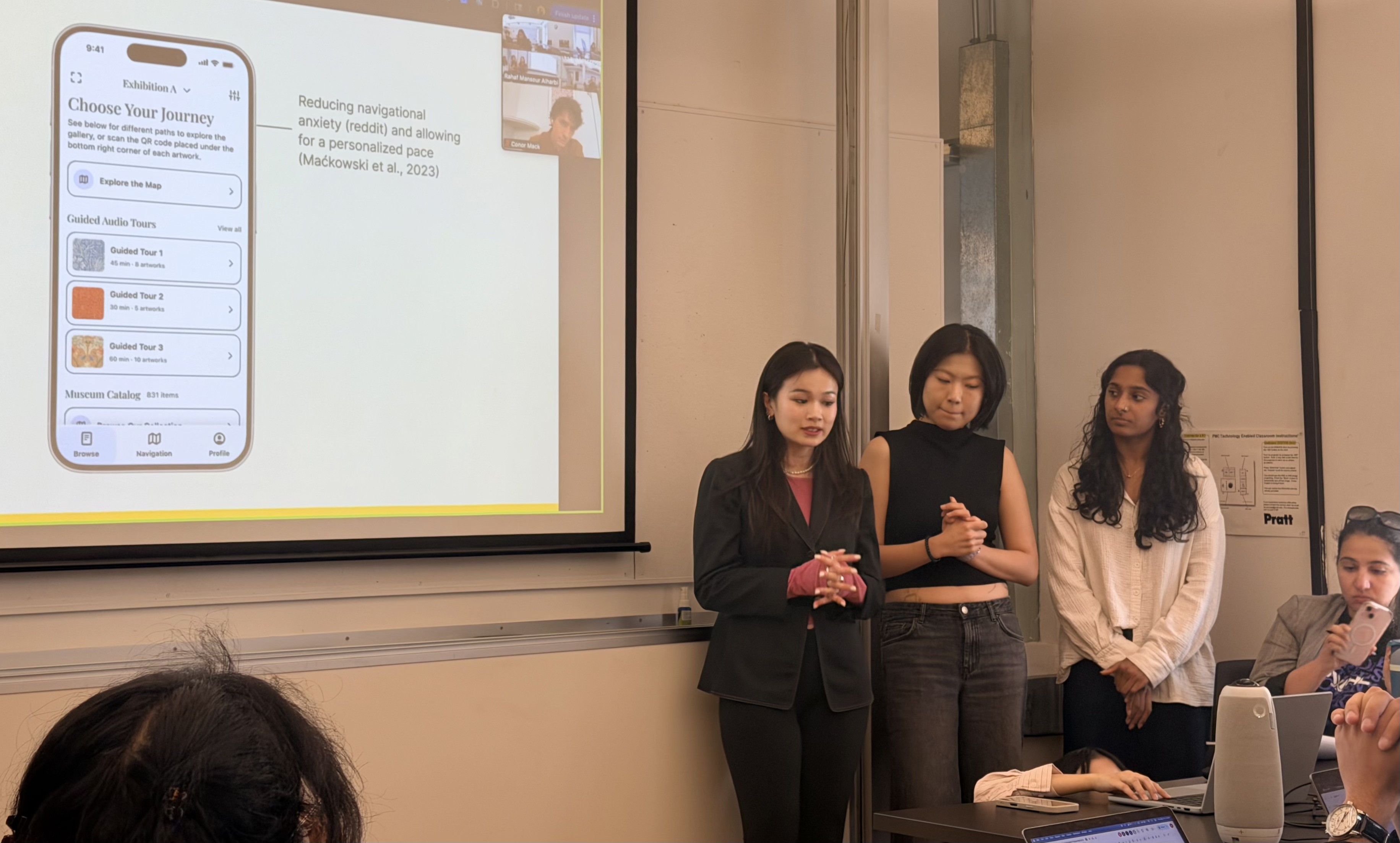 Three people stand presenting in a classroom, showing a slide about a mobile app for museum tours. The mood is focused and professional.