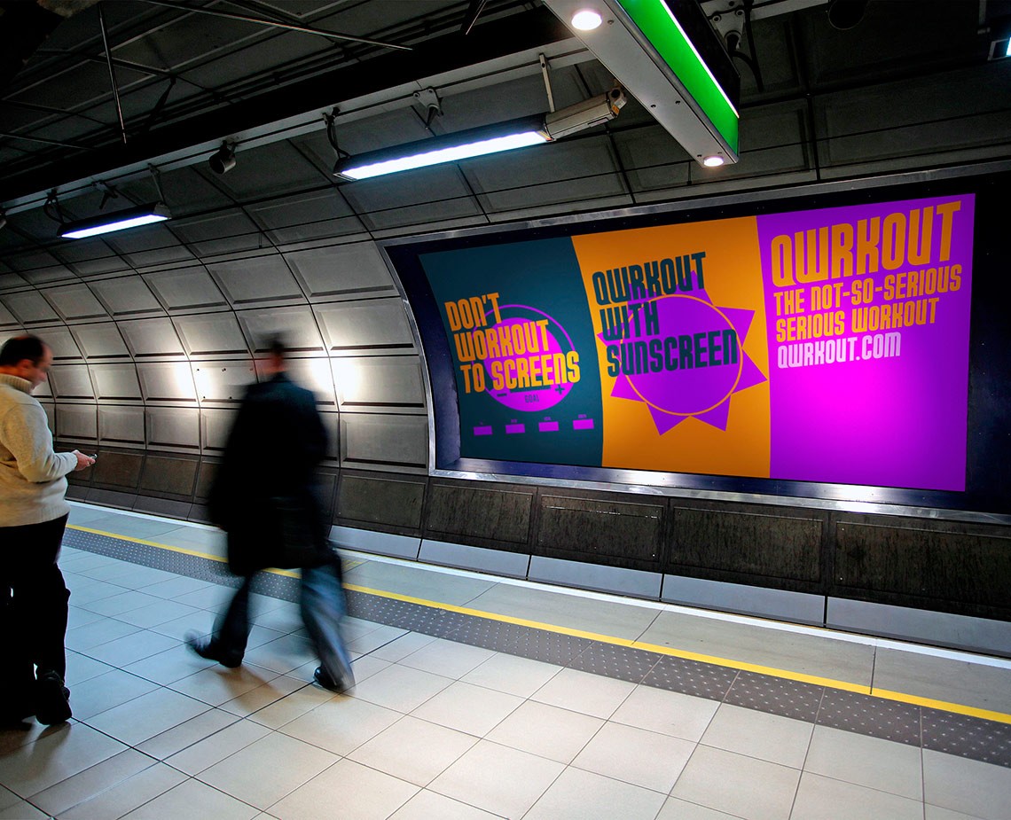 Two people are walking on a subway platform beneath a large advertisement. The advertisement features bright colors and text reading “DON’T WORKOUT TO SCREENS” and “QUIRKOUT THE NOT-SO-SERIOUS SERIOUS WORKOUT.QUIRKOUT.COM”. The platform has white tiles and a yellow line along the edge. The ceiling of the subway station is dark with visible metal beams and fluorescent lighting.