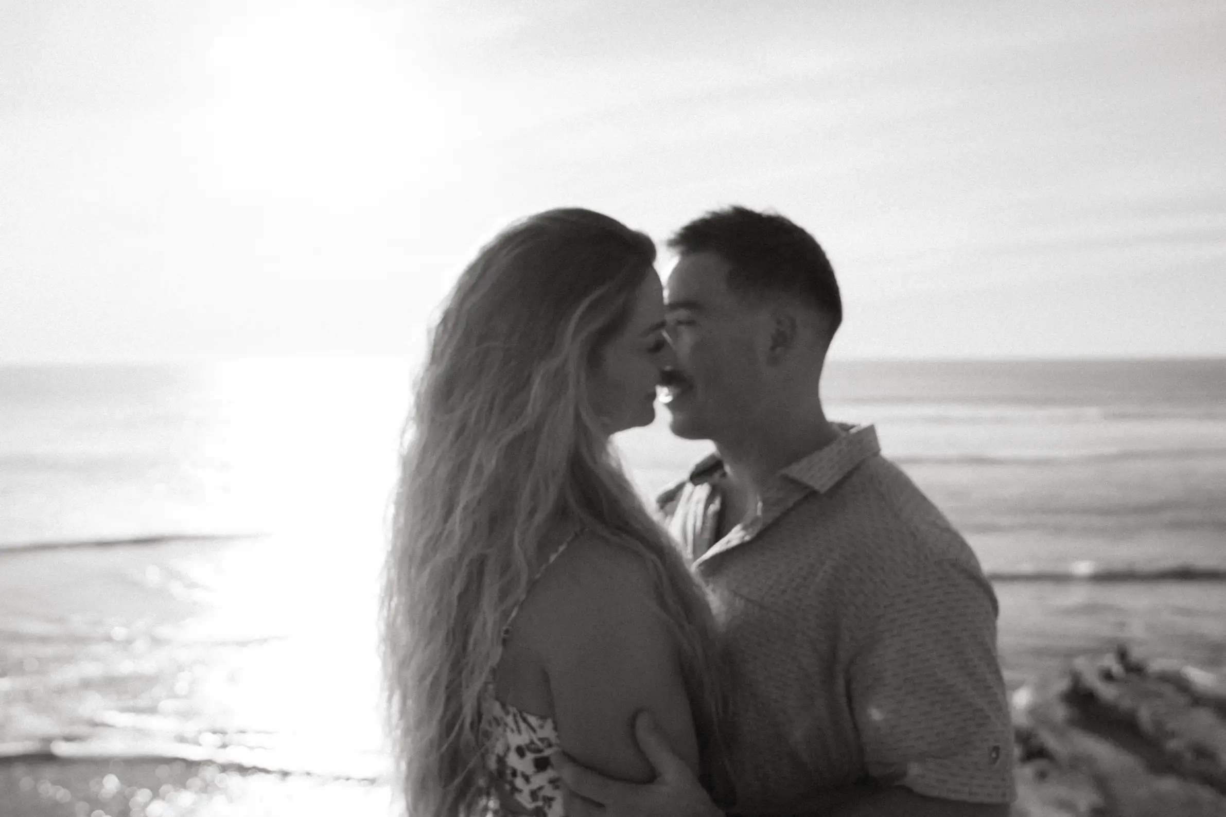 A black and white photo of a couple embracing close to the ocean, silhouetted against the light.