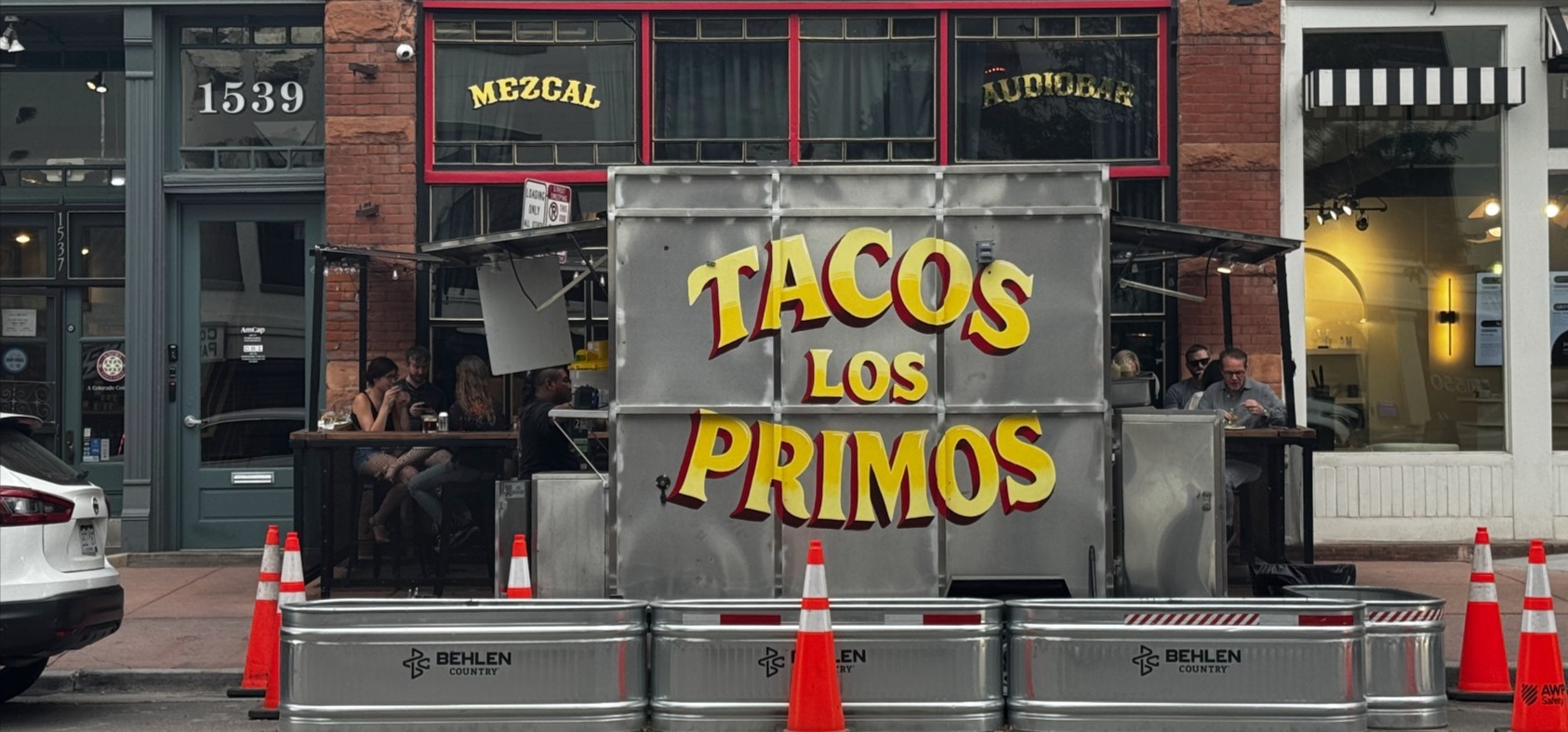 A food truck with the name "Tacos Los Primos" painted on the side, in front of the Mezcal Audiobar on Platte Street in Denver.