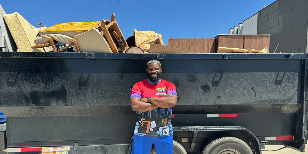 A man stands confidently in front of a large, filled dumpster under a clear blue sky. He's wearing a colorful shirt and blue shorts, smiling proudly.