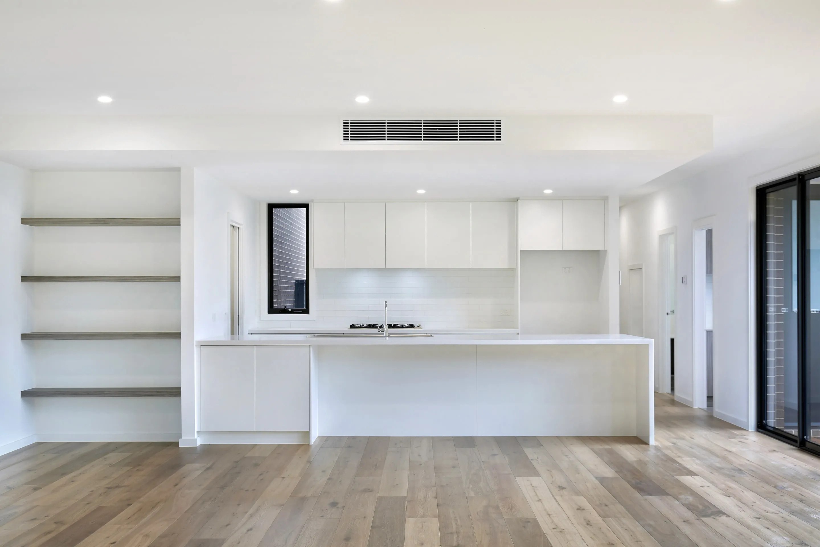 Open living and kitchen with white walls, oak floors, grey floating shelves, a white kitchen with island and gas cooktop under a black-framed window, and a black sliding door to the side