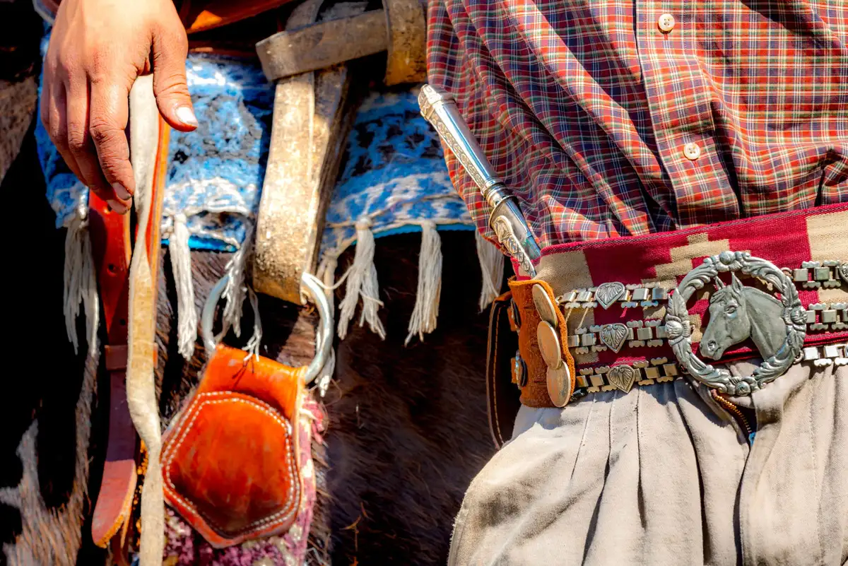 A person in a plaid shirt and traditional white trousers holds a wooden staff next to a decorated horse's head.