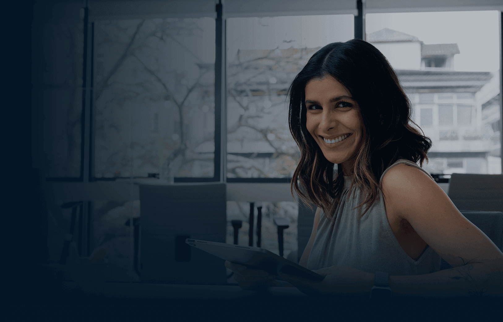 Uma mulher de blusa amarela olha para o celular, sentada perto de uma janela em uma sala suavemente iluminada.