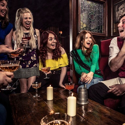 Group of five people laughing and holding drinks around a table with candles in a cozy, dimly-lit room with stained glass windows.
