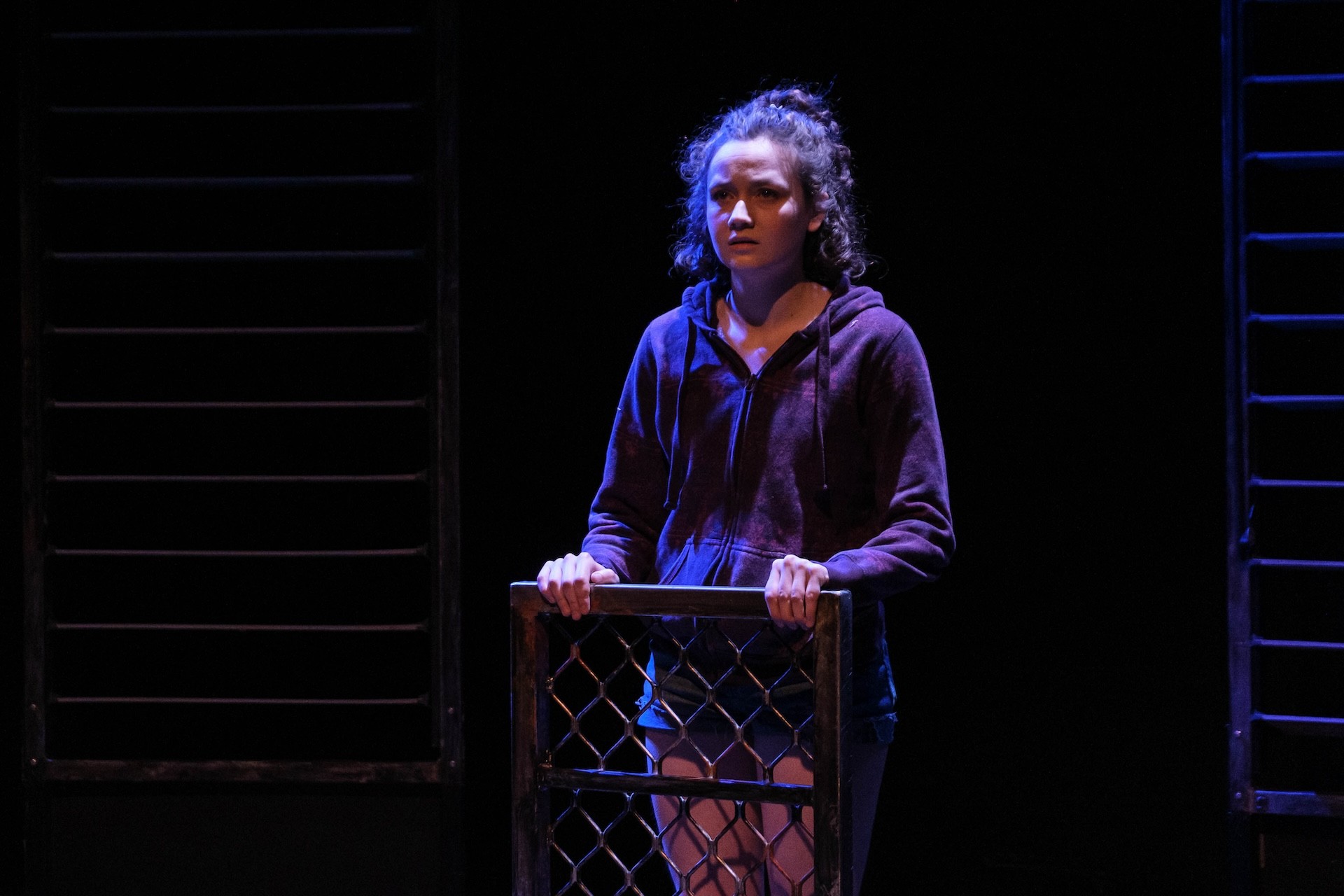 A performer in a hoodie grips the top of a metal barrier, looking tense and distressed as cool blue stage lighting highlights her expression.