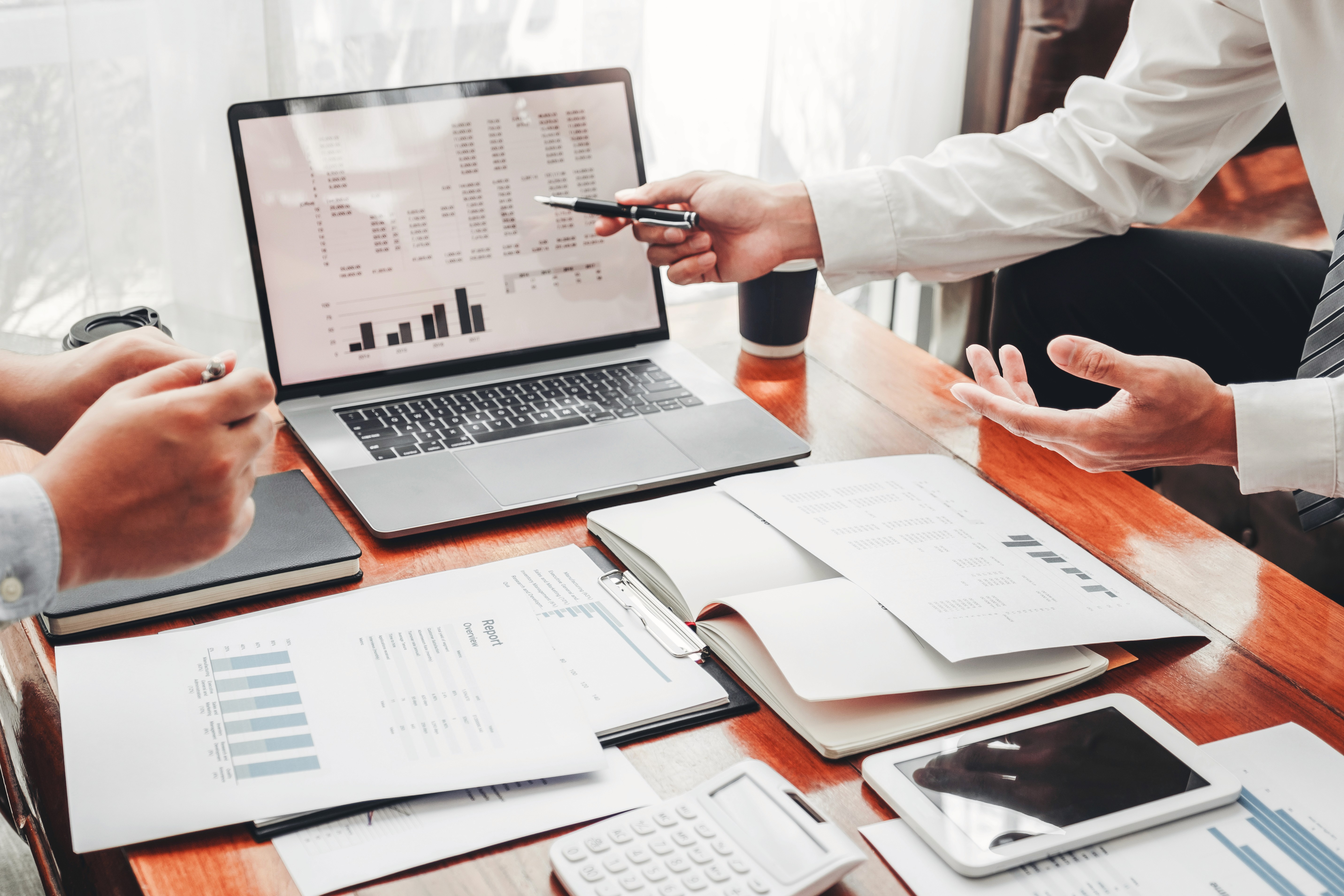 Two people are engaged in a business meeting, with financial charts and graphs displayed on a laptop screen, papers, and a digital tablet spread across a wooden table, highlighting a collaborative work environment.
