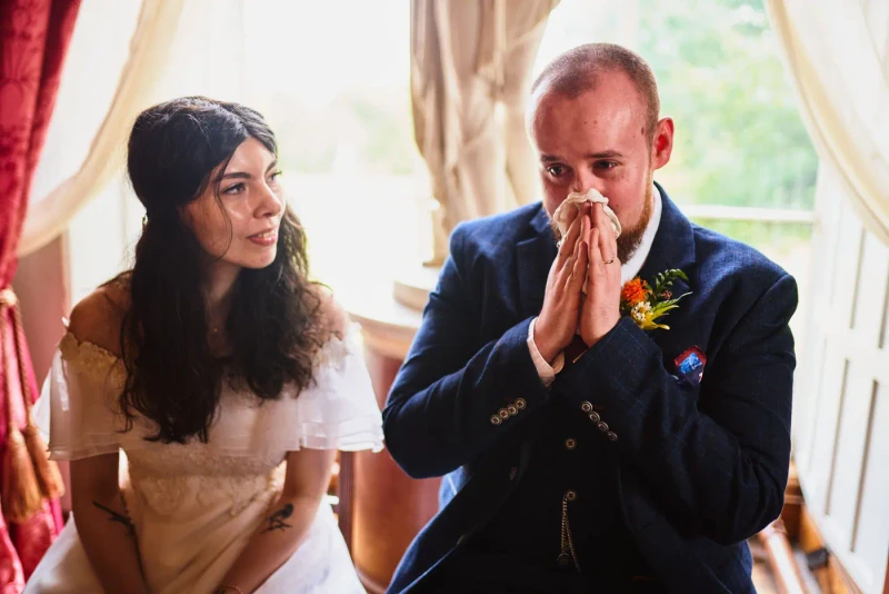 Groom with his hands pressed to his face, overcome during the ceremony as his bride smiles
