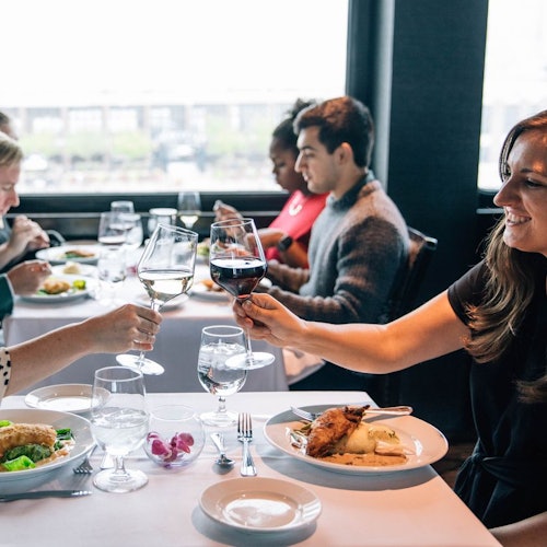 Mga tao na nakaupo sa isang hapag-kainan sa tabi ng bintana, nagtutustusan gamit ang mga baso ng alak, may mga plato ng pagkain at kubyertos sa harapan nila.