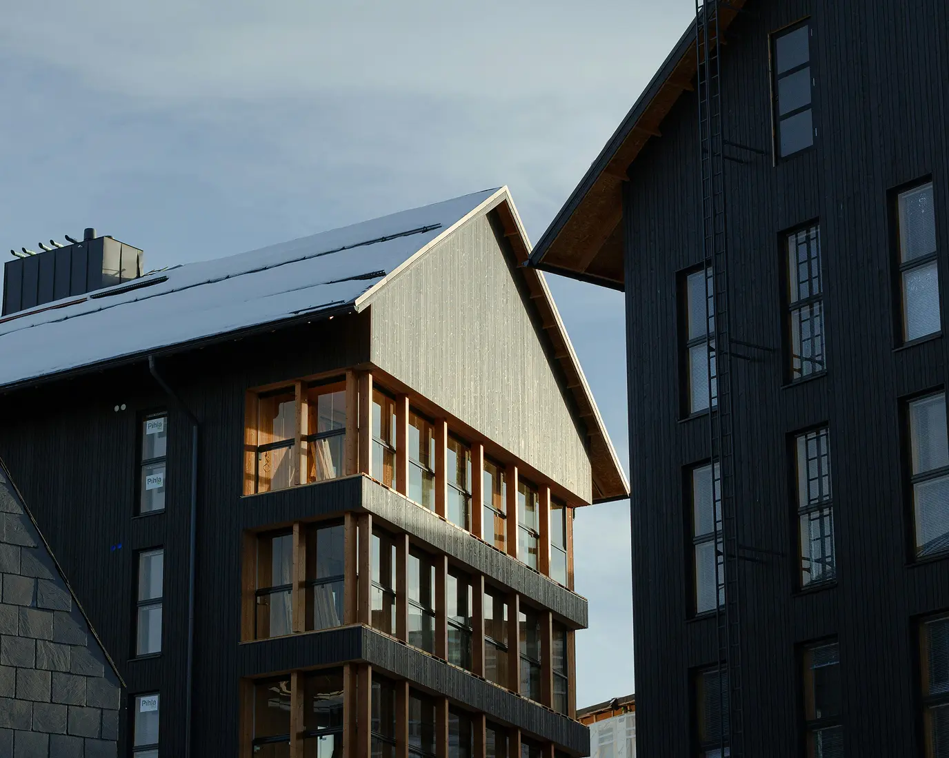 Modern buildings with dark wooden facades and large windows under a cloudy sky. The sunlight casts a soft glow, creating a serene, minimalist tone.