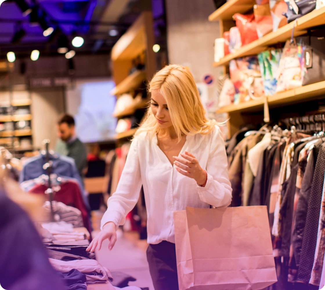 A woman shopping in a boutique, browsing through clothes on a rack with a pink shopping bag in hand.