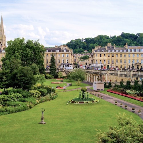 一个精心修剪的公园,绿草如茵,花团锦簇,中央设有喷泉,四周被历史建筑和树木环绕,天空中有些许白云点缀。