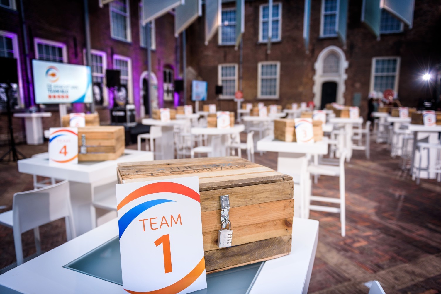Tafel met een houten kist met cijferslot en een bordje met de tekst ‘Team 1’ in een zaal met meerdere opgestelde tafels en kisten.