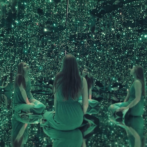 一位穿绿色衣裙的女士盘腿坐在一个充满绿色灯光的镜室里,创造出多个倒影。