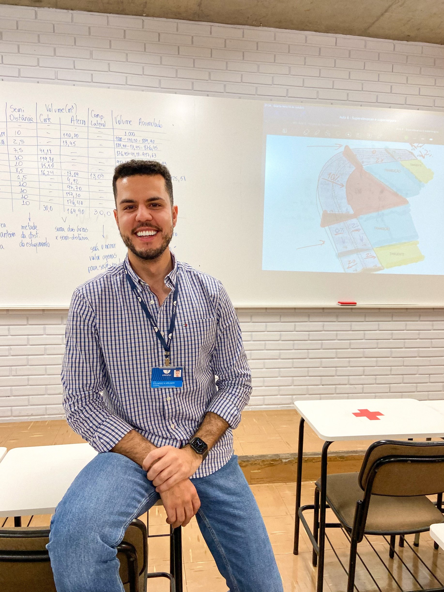 Professor Mestre Eduardo Alexandre Kruger sorrindo em sala de aula com quadro branco contendo tabelas de volumes de corte, aterro e curvas de nível.
