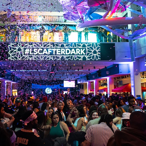Una gran multitud se reúne bajo luces coloridas mientras cae confeti. Un letrero en el fondo dice "#LSCAFERDARK".