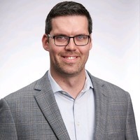 A man with glasses and a light gray blazer smiles against a softly lit background.