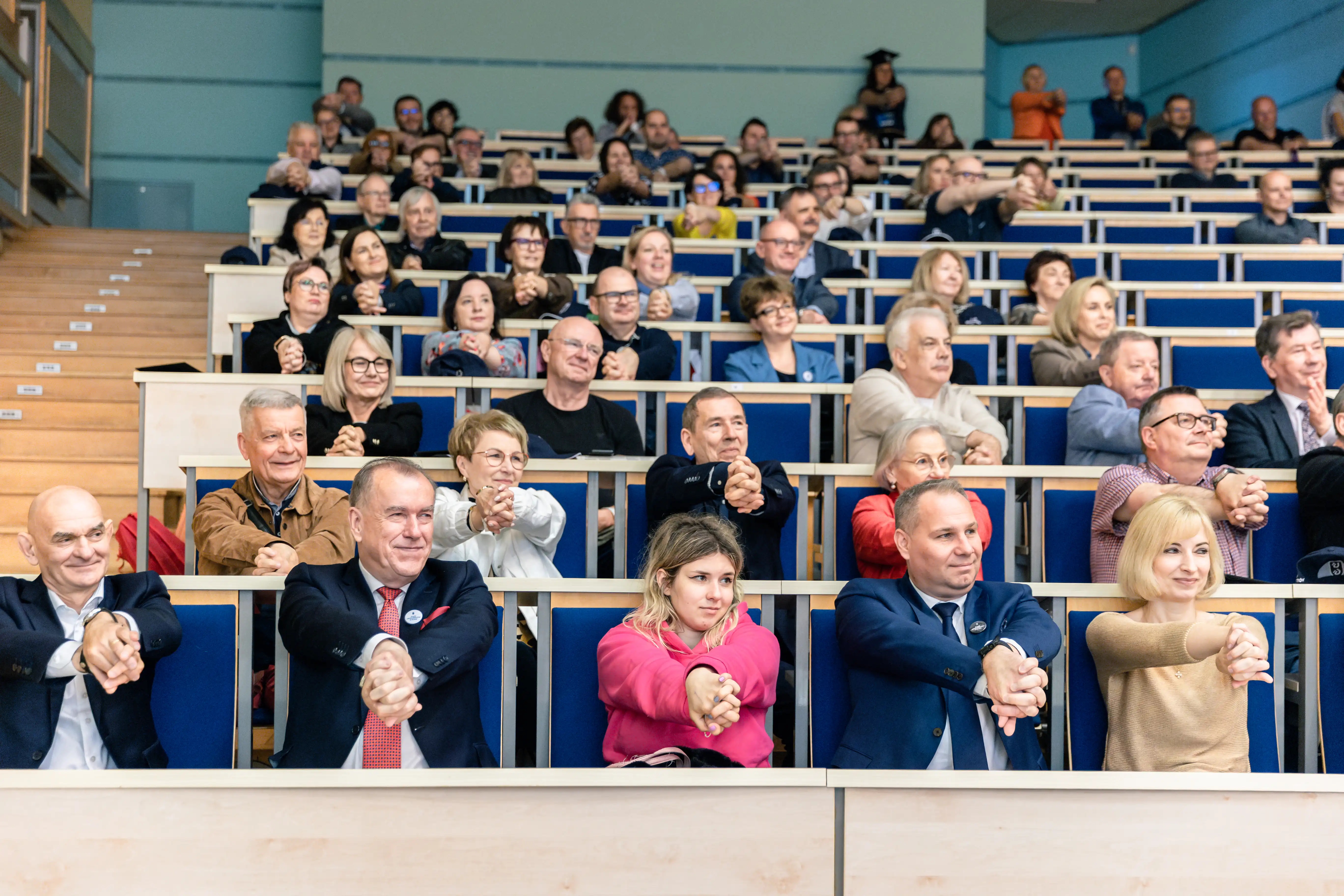 Grupa ludzi siedząca na auli uczelnianej. Ubrani w eleganckie stroje, wszyscy wykonują podobny gest rękoma, wyciągniętymi do przodu.