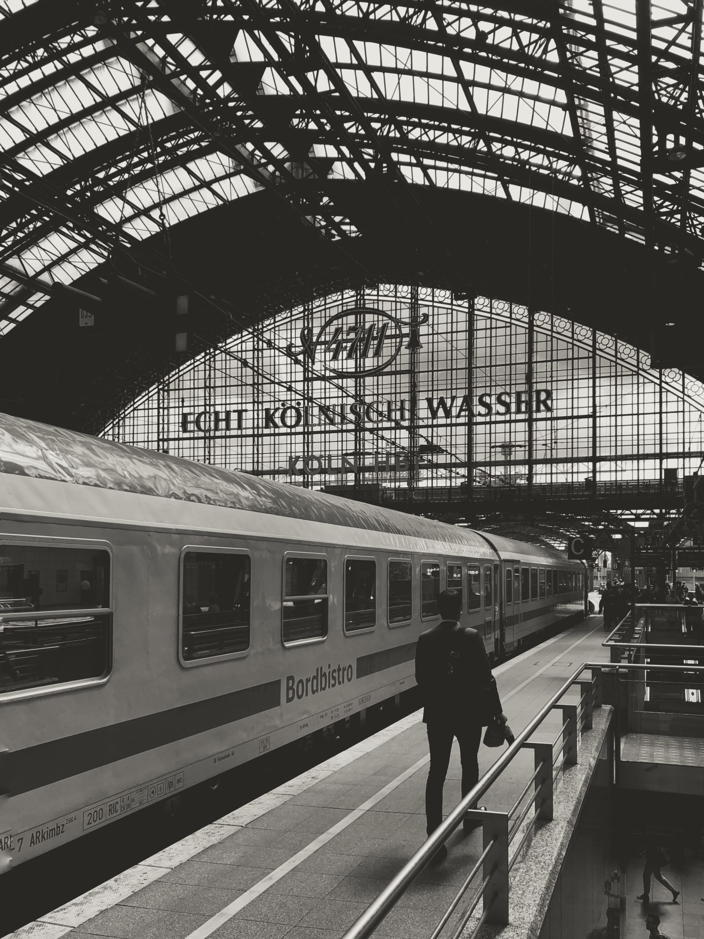 Black-and-white travel photo of Cologne Central Station (Köln Hauptbahnhof) highlighting historic train station architecture, the Bordbistro carriage, and the iconic 4711 Cologne advertisement — ideal for content about train travel, Cologne landmarks, and architectural photography.