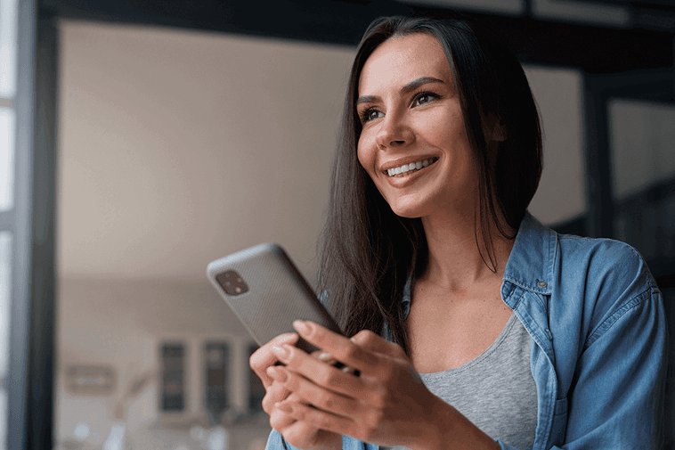 Smiling woman holding a smartphone indoors, looking slightly to the side. She wears a casual outfit with a denim shirt and gray top, appearing happy and engaged with her mobile device, representing connection, communication, and digital interaction.