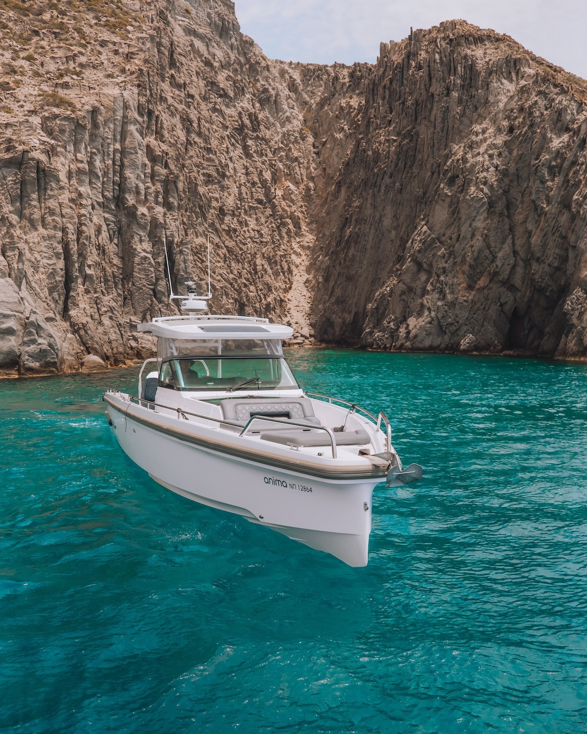 White Axopar 37 yacht anchored in turquoise waters near dramatic limestone cliffs and natural rock arch in the Cyclades.