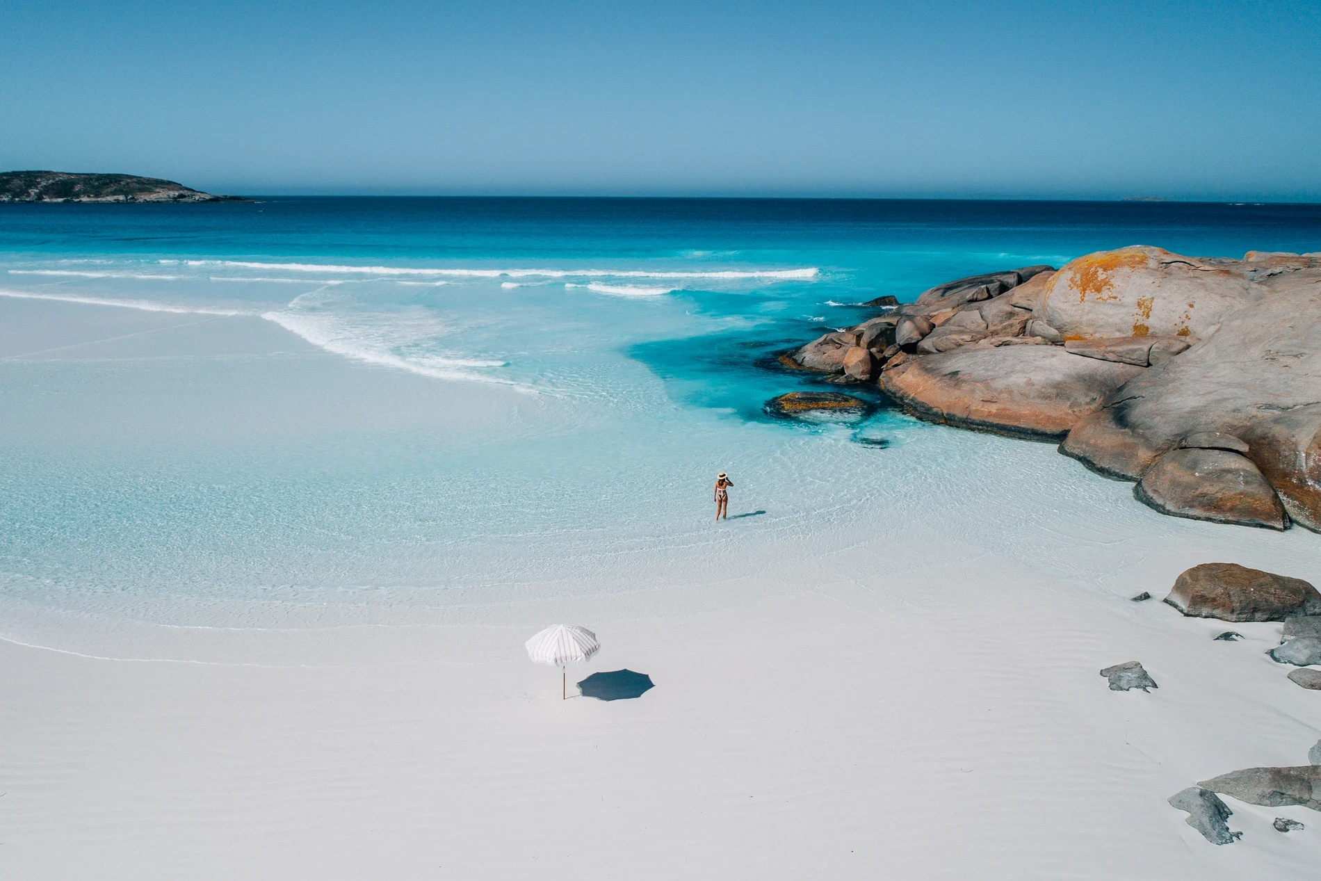 A white umbrella on white sand beside granite boulders and clear turquoise water