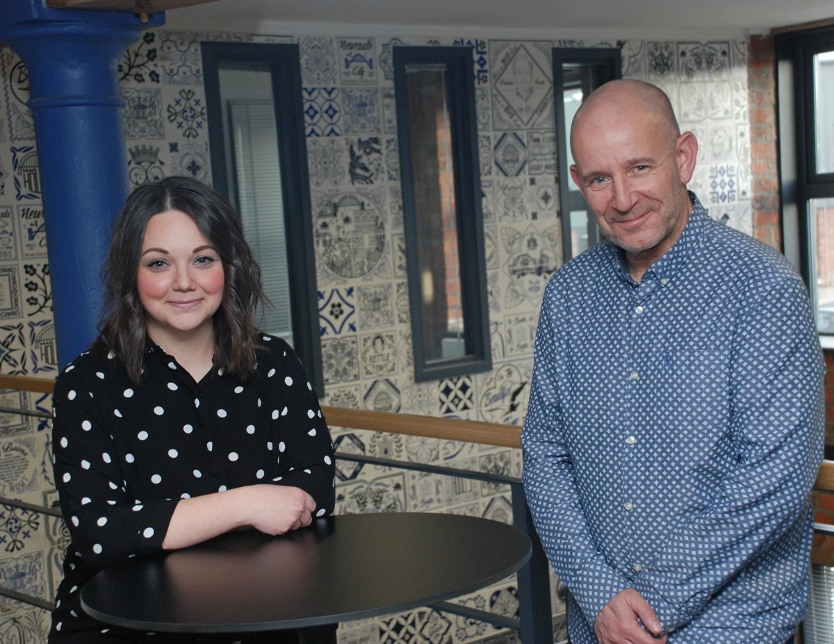 Two people stand beside a tall round table in an indoor space with patterned tile artwork and narrow windows on the wall behind them.