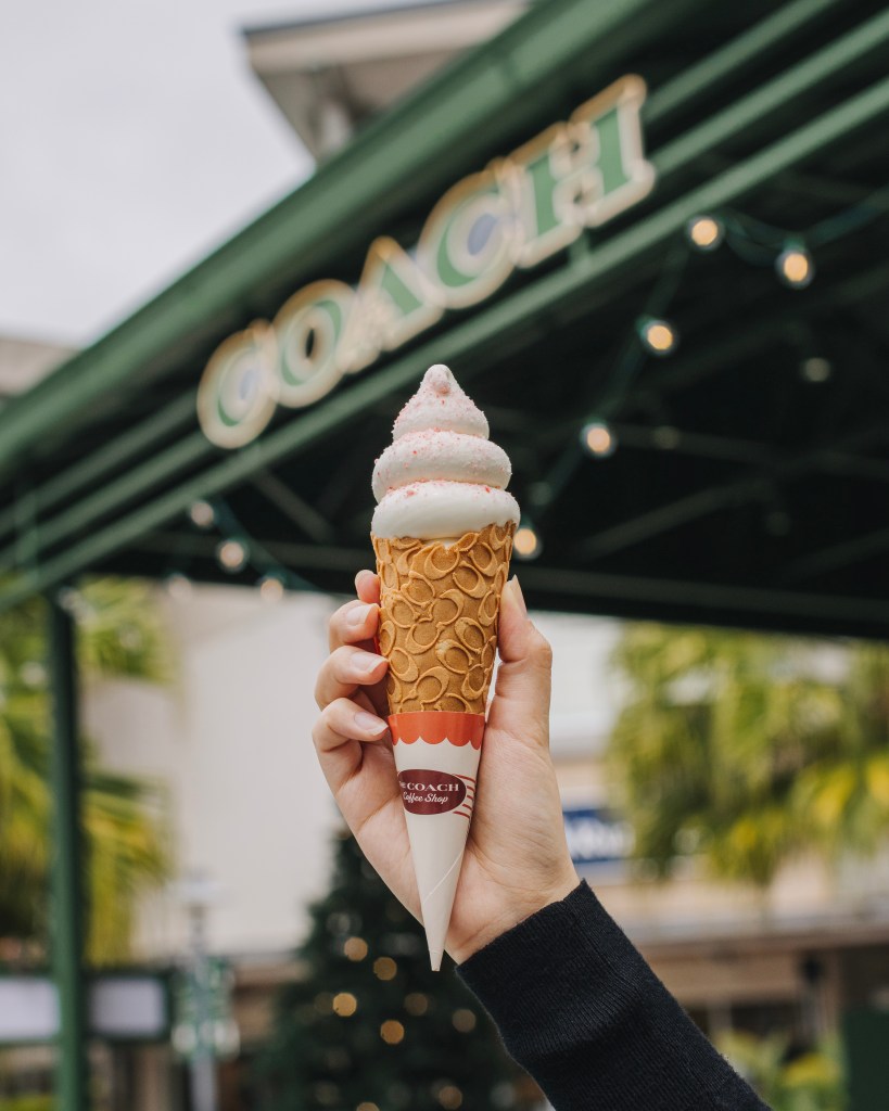 A hand holding a cone of soft-serve ice cream in front of a Coach pop-up shop decorated with festive lights.