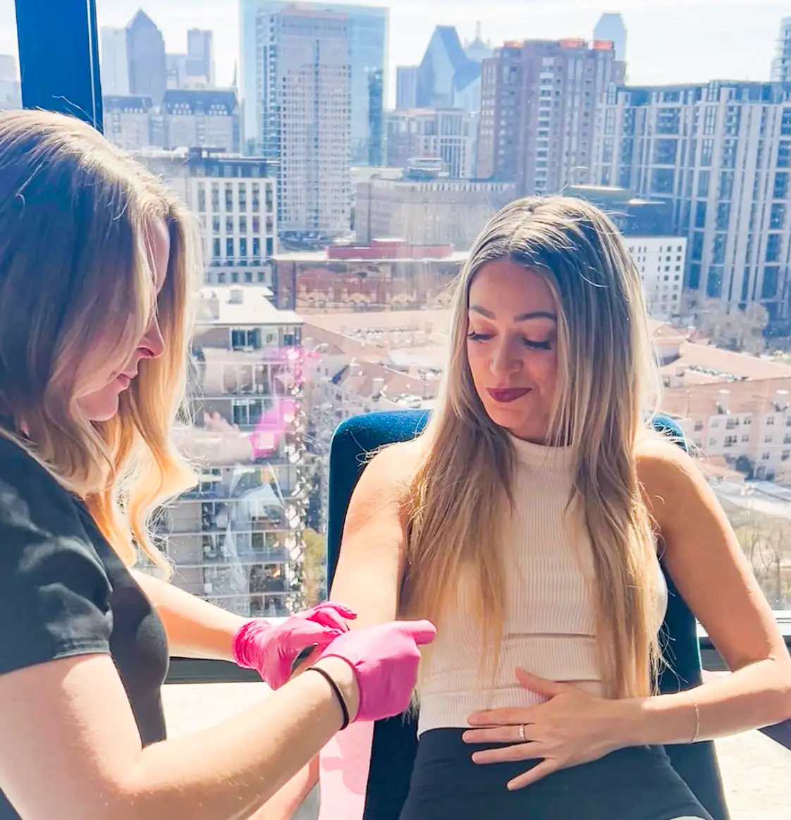 An Event Hydration nurse in pink gloves administering an IV drip to a smiling woman seated in front of a window overlooking the Dallas skyline