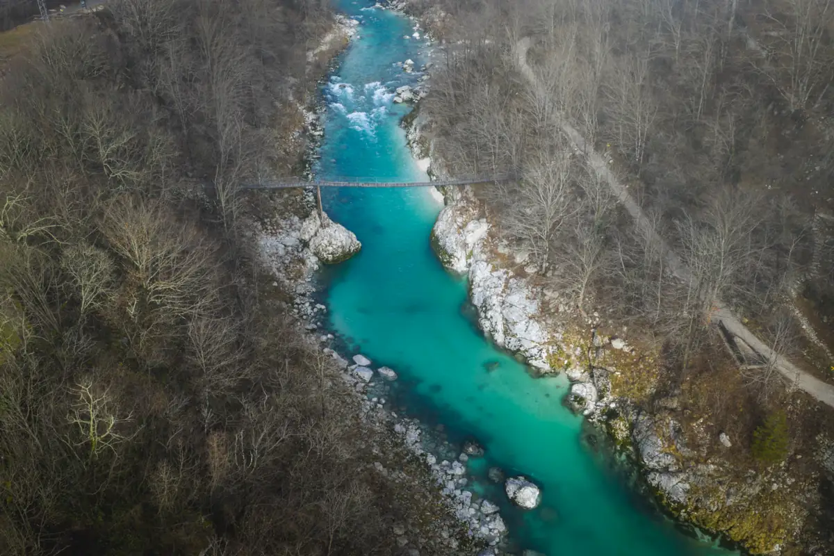  Aerial view of the emerald-colored Soča River flowing through a rocky gorge in the Soča Valley, Slovenia, featuring a narrow pedestrian suspension bridge over turquoise waters and leafless trees during early spring.