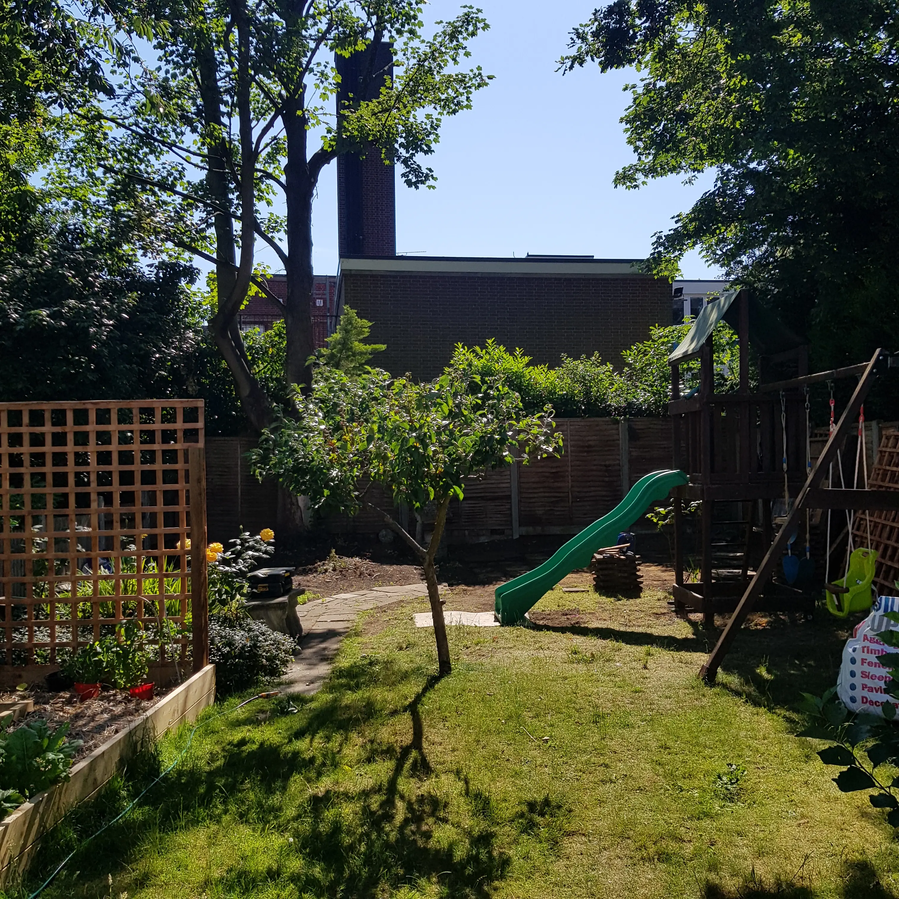 A sunny garden featuring greenery, a wooden trellis, a slide, and a distant building in the background.