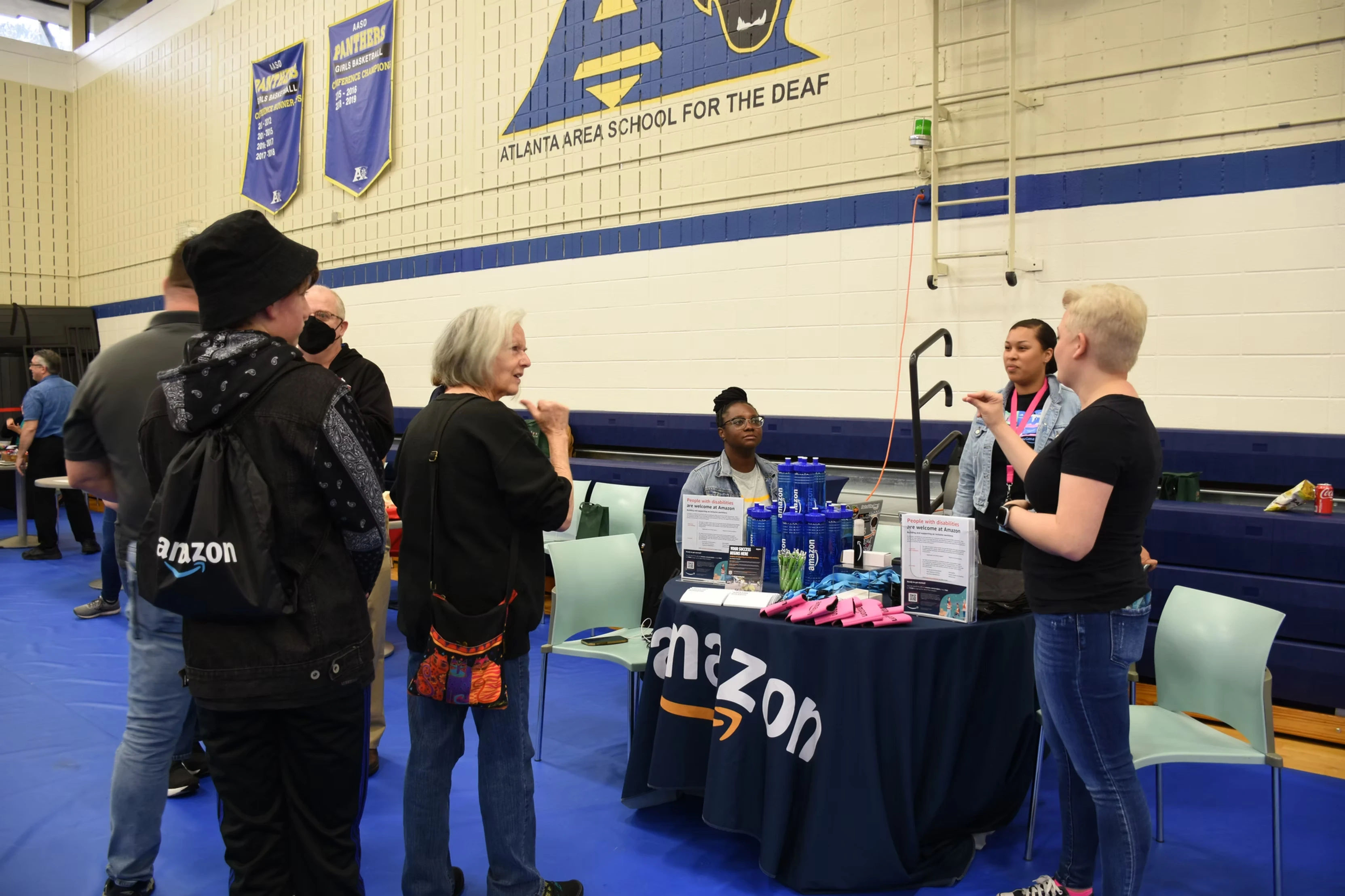 Amazon representatives talk with students at a career fair. A table displays Amazon branding and products.