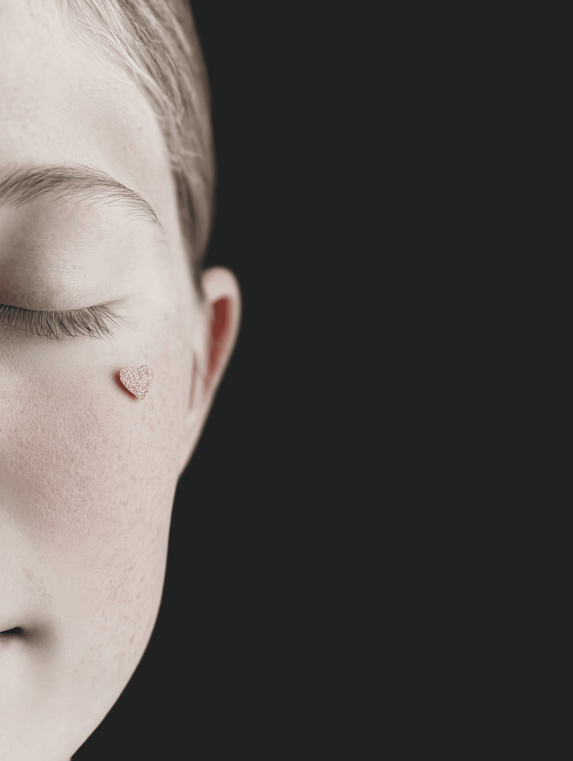 Close-up portrait of a person with eyes closed, featuring a small pink heart sticker on the cheek against a dark background.