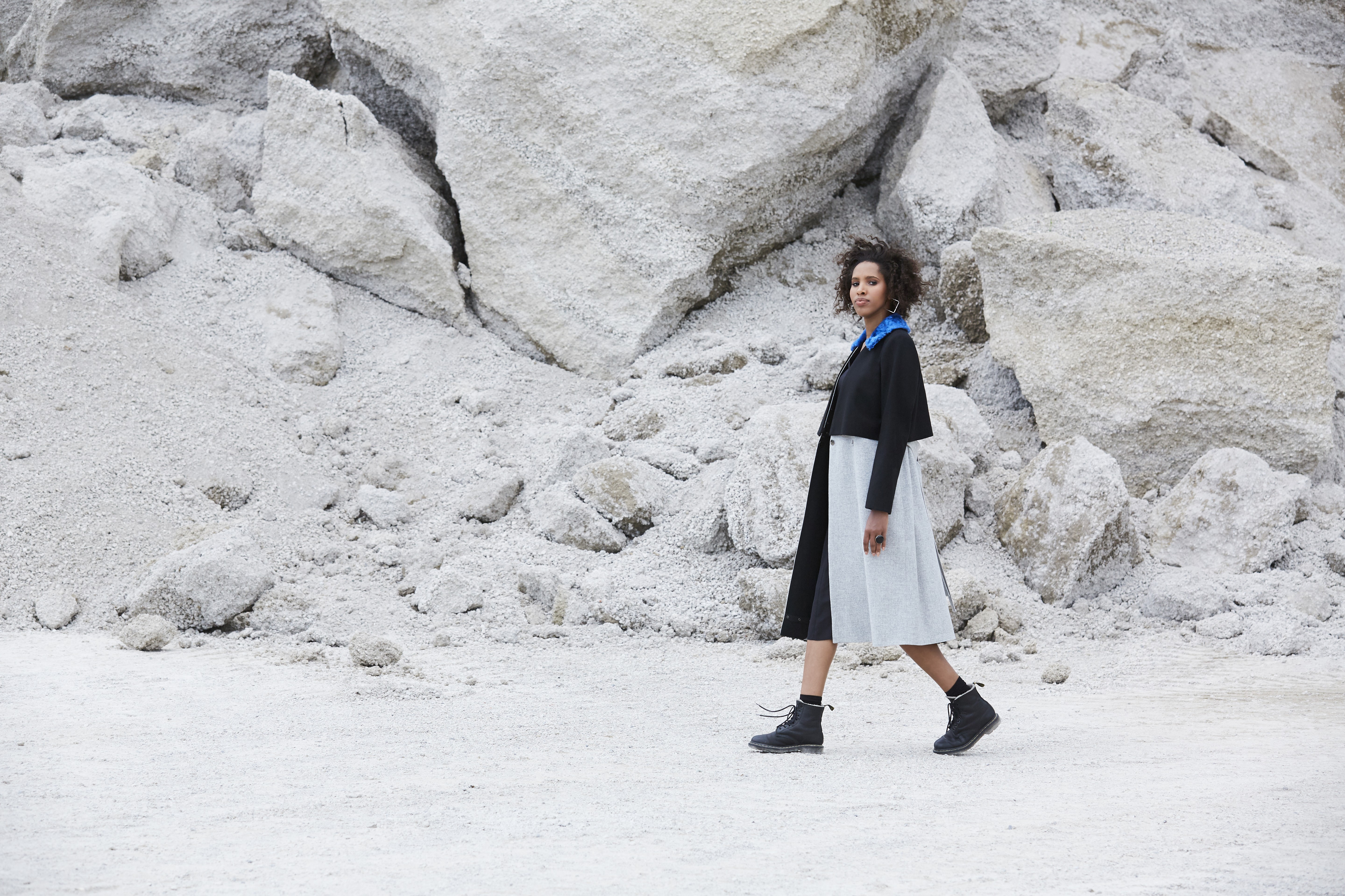 A person wearing a stylish black and gray coat with blue accents walks across a rugged landscape of large, textured white rocks and gravel.